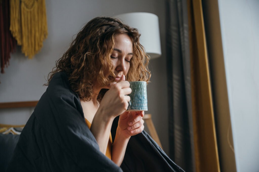 A young woman inhales the aroma of hot coffee in the early morning while sitting in bed. Beautiful middle aged girl with mug in bed with closed eyes Relaxing cannabis drink before bed